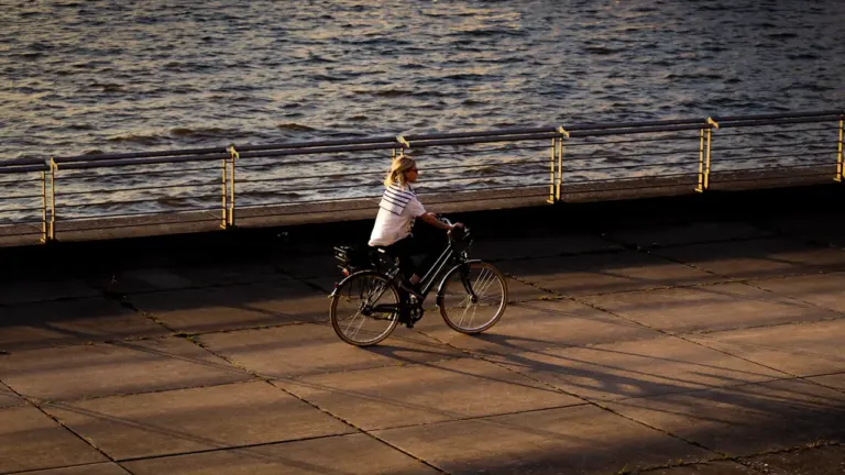 Eine Radfahrerin fährt bei sonnigem Wetter an einem Flussufer entlang in Deutschland.