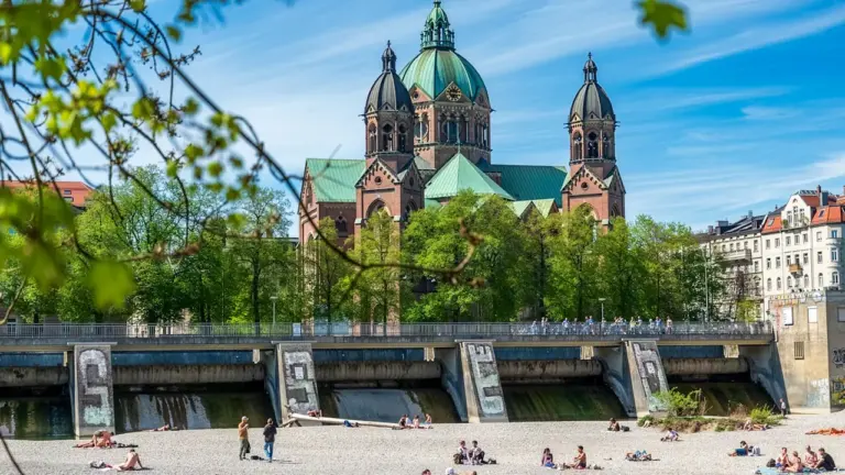 Menschen entspannen sich an einem sonnigen Sommertag am Isarufer in München vor einer Kirche.