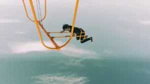 Eine Person hängt in luftiger Höhe an einem gelben Metallrahmen über den Wolken.